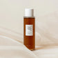 A brown bottle of 'Beauty of Joseon Ginseng Essence Water' with a white cap, placed on a textured fabric, likely a linen fabric.