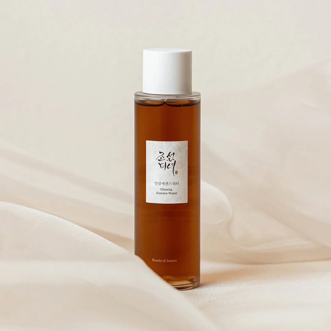 A brown bottle of 'Beauty of Joseon Ginseng Essence Water' with a white cap, placed on a textured fabric, likely a linen fabric.